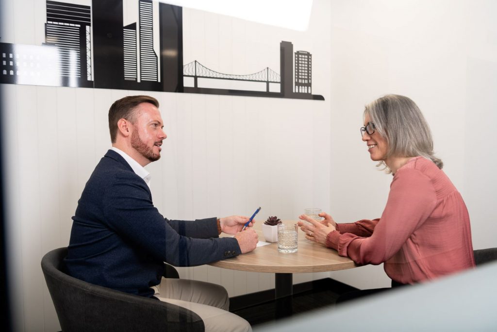 A man in a suit jacket and women in a pink shirt sit in a small office and talk across a table about how they can prepare for a job interview.