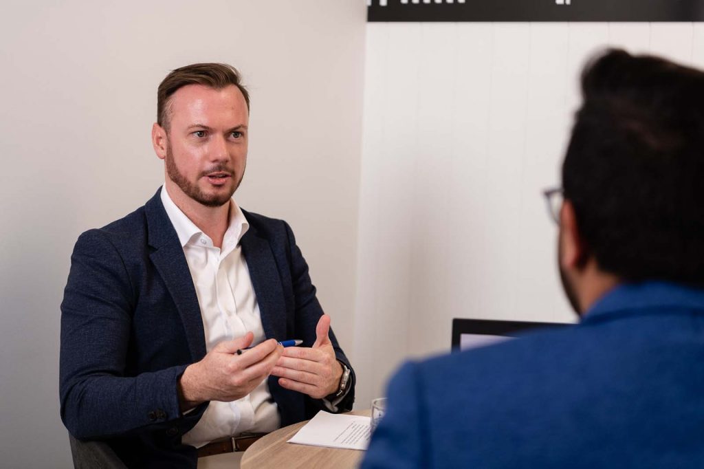 An IT leader talks to an Exclaim IT Connections partner about an executive role. They are sitting in a small office. The recruiter is wearing a blue suit and has his back to the camera. The candidate is wearing a navy jacket and white button up shirt and gesturing with his hands as he talks about his skillset and experience.