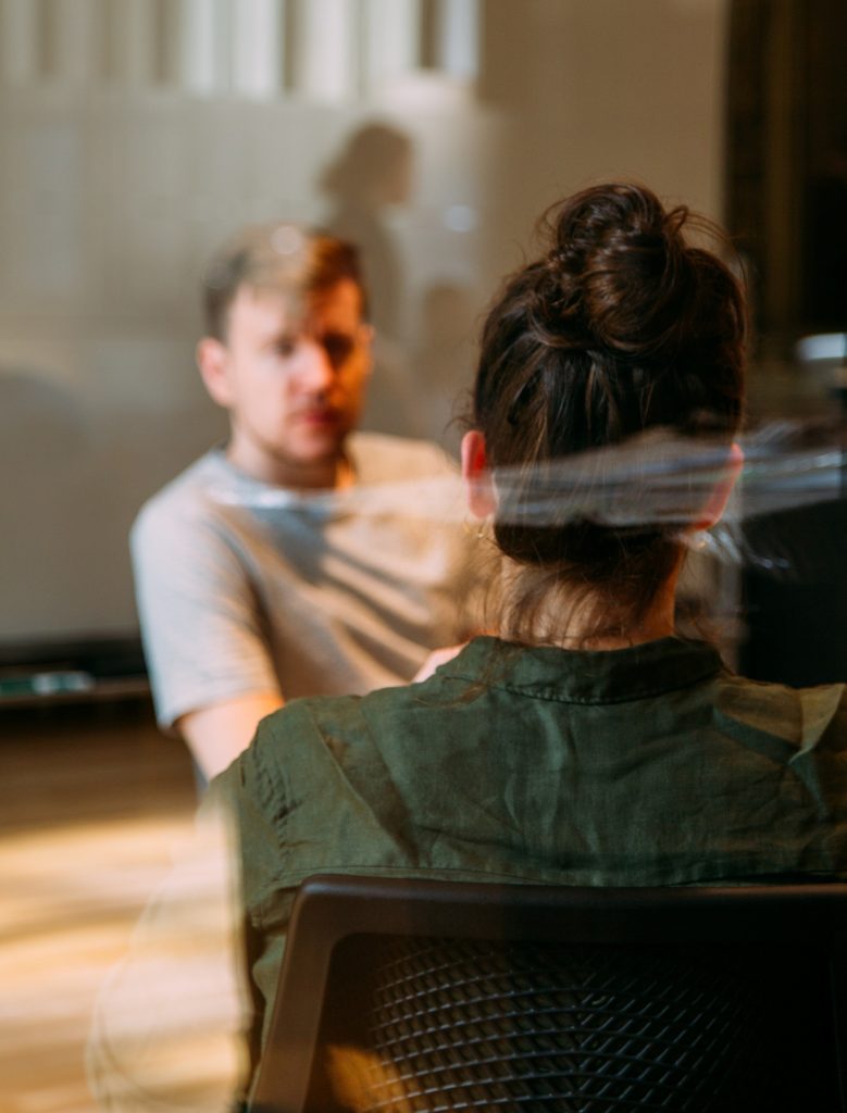 Man and woman discuss executive search requirements. Woman is wearing a green long-sleeved shirt and has her back to the camera. Man looks at her across the table and is blurred. He wears a grey shirt.